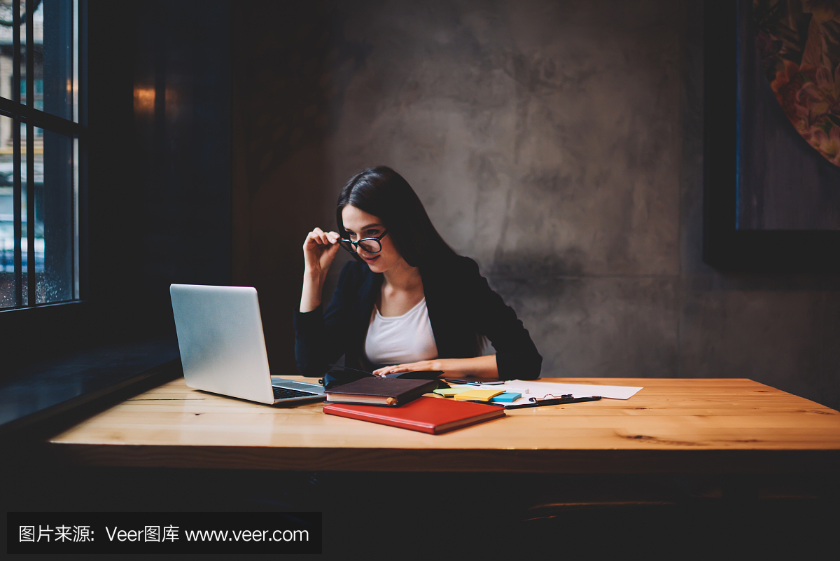 专注的年轻女子,戴着时尚的眼镜,穿着正式的黑色夹克,坐在木制的桌子前,用现代的上网本设备在网上观看视频
